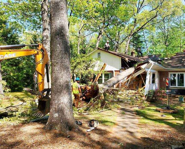 Excavator lifting a storm-fallen tree off a damaged house